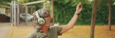 Panorama, orta yaşlı, dost canlısı, gri saçlı ve sakallı sıradan kıyafetler giyen kulaklıkla müzik dinleyen bir adam. Gözlüklü, olgun bir beyefendi açık havada müzik dinlemekten hoşlanır.
