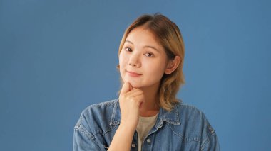 Kot gömlek giymiş, bir şeyler düşünen, iyi bir fikri olan, stüdyoda mavi arka planda izole edilmiş genç bir kadının yakın çekimi.
