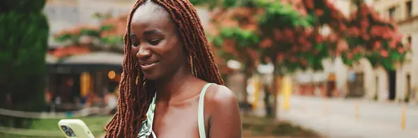 Uśmiechnięta Kobieta Afrykańskimi Warkoczami Nosząca Top Stoi Ulicy Używa Telefonu — Zdjęcie stockowe