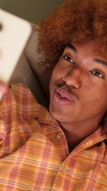 Gülümseyen adam oturma odasında yatarken cep telefonuyla video görüşmesi yapıyor.