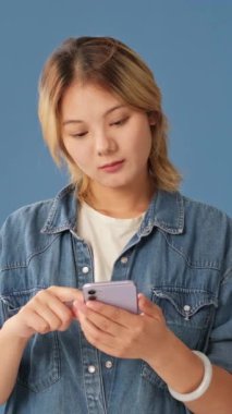 Şaşırmış genç bir kadın mavi arka planda izole edilmiş cep telefonu kullanarak iyi haberler alıyor.