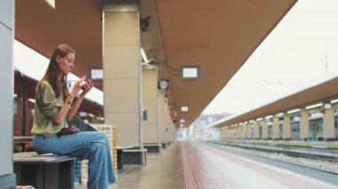 Genç bir kadın, tren istasyonundaki bankta otururken cep telefonuyla sesli mesaj gönderiyor.
