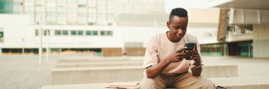 Genç Afrikalı öğrenci üniversitenin dışında oturuyor, cep telefonu kullanıyor, akıllı telefon ekranını dinliyor, Panorama