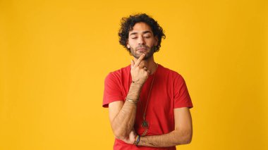 Dalgın, kıvırcık saçlı, kırmızı tişörtlü, çenesini ovuşturan ve stüdyoda sarı arka planda yaşayan çekici bir adam.