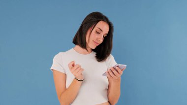 Beyaz elbiseli güzel esmer kadın cep telefonu parşömenleri kullanıyor. Stüdyoda mavi arka planda izole edilmiş online alışveriş yapıyor.