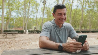 Gri tişört giyen orta yaşlı bir adam park bankında otururken cep telefonu kullanıyor.