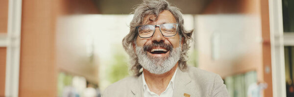 Mature businessman with beard in eyeglasses wearing gray jacket sits in cafe, Panorama. Middle aged manager having video conference, video call on laptop pc computer sit at cafe outdoor
