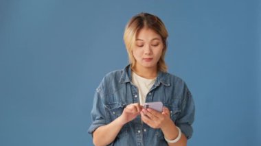 İnternetten alışveriş yapan genç bir kadın mavi arka planda izole edilmiş cep telefonu kullanıyor.