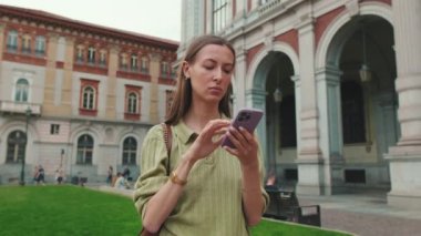 Güzel bir kadın eski kasaba meydanında dururken cep telefonunda harita uygulaması kullanıyor.