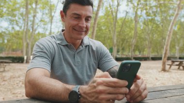 Gri tişört giyen orta yaşlı bir adam park bankında otururken cep telefonu kullanıyor.