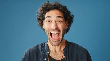 Yaklaş, kıvırcık saçlı, mavi gömlekli, kollarını kavuşturan ve stüdyoda mavi arka planda izole edilmiş kameraya bakan çekici bir adam.