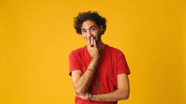 Kıvırcık saçlı, kırmızı tişörtlü hoşnutsuz bir adam. Stüdyoda sarı arka planda izole edilmiş bir kameraya bakarken anlaşmazlık gösteriyor.