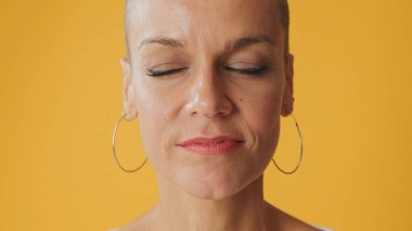 Kılsız genç bir kadının portresi gözleri kapalı stüdyoda sarı arka planda duruyor.