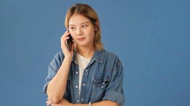 Kot gömlek giymiş genç bir kadın cep telefonuyla konuşuyor. Stüdyoda mavi arka planda izole bir şekilde kazanıyor.