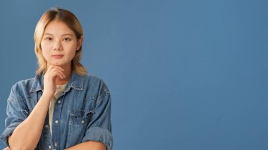 Kot gömlek giymiş genç bir kadın kameraya özenle bakıyor. Model mekan, stüdyoda mavi arka planda izole edilmiş.