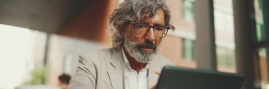 Panorama, gözlüklü sakallı, gri ceketli, tablet üzerinde çalışan, açık hava kafede oturan olgun bir iş adamı. Başarılı bir adam doğadaki kafede oturuyor. Mobil ofis çalışanı.