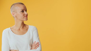 Kılsız genç bir kadın, kollarını kavuşturmuş ve gülümsüyor. Stüdyoda sarı arka planda izole edilmiş bir yer arıyor.