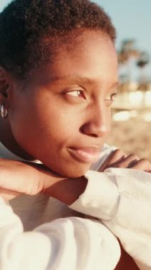 Açık renkli günlük elbiseli mutlu kız sahilde oturuyor, gülümsüyor ve uzaklara bakıyor.