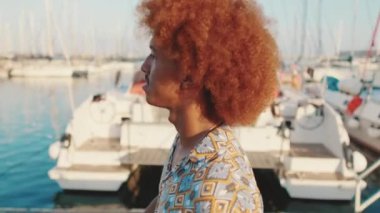 Yazın beyaz yatların arka planında yürüyen ve gülümseyen kıvırcık saçlı genç adamın yan görüntüsü.