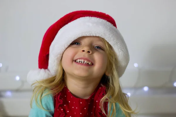 Kırmızı Noel Baba şapkalı küçük sarışın kız ve arka plandaki Noel süsü kameraya bakıyor ve gülümsüyor, yeni yıl kostümlü çocuk portresi, kış tatili resmi..