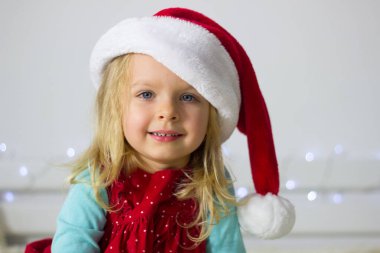 Kırmızı Noel Baba şapkalı küçük sarışın kız ve arka plandaki Noel Baba süslemesi kameraya bakıyor ve gülümsüyor, yeni yıl kostümlü çocuk portresi, kış tatili resmi..