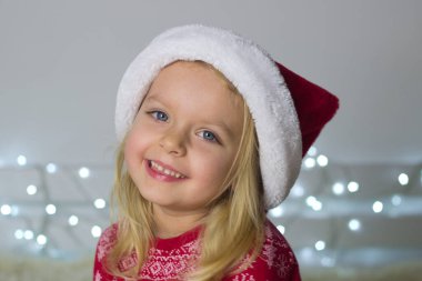 Little blond girl in red Santa hat and Christmas decoration on background and smile,portrait of kid in New year pyjama,winter holidays image.