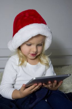 Kırmızı Noel Baba şapkalı küçük sarışın kız elinde tablet, elinde bebek elinde aletle internet kullanıyor tablet ve çizgi film bağımlısı çocuk, gelişim oyunlarıyla internetteki çocuk.