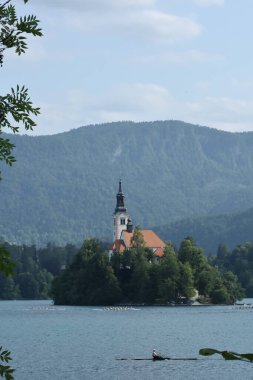 Bled Island 'daki ikonik kilise ön planda kürekçiler ve arka planda yeşil dağlar..