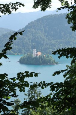 Adanın ikonik kilisesiyle Bled Gölü manzarası, doğal olarak ağaç dalları tarafından çerçevelenmiş..