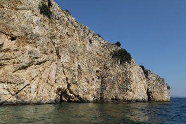 Engebeli bir kireçtaşı kayalığı berrak mavi suyun üzerinde yükseliyor. Parlak, güneşli bir günde yakalanan bu uzak kıyı manzarasının dokuları ve gölgeleri doğal güzelliğini arttırır..