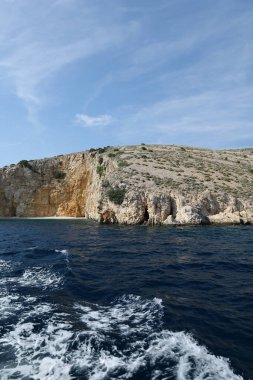 Engebeli bir kireçtaşı kayalığı berrak mavi suyun üzerinde yükseliyor. Parlak, güneşli bir günde yakalanan bu uzak kıyı manzarasının dokuları ve gölgeleri doğal güzelliğini arttırır..
