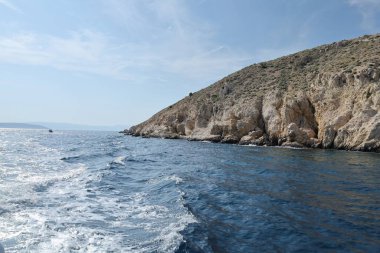 Engebeli bir kireçtaşı kayalığı berrak mavi suyun üzerinde yükseliyor. Parlak, güneşli bir günde yakalanan bu uzak kıyı manzarasının dokuları ve gölgeleri doğal güzelliğini arttırır..