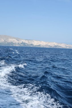 Dalgalar ön planda dalgalanıyor ve kırılıyor. Uzaktaki dağlar sabah sisi altında hafifçe yükselirken..