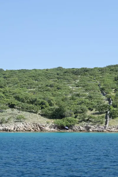 Kayalık bir sahil şeridindeki yeşil bir deniz feneri direğinin yanında yıpranmış bir taş ev duruyor. Engebeli manzara ve parlak mavi gökyüzü doğa ve unutulmuş insan yapıları arasındaki zıtlığı vurgular..