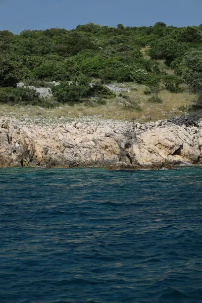 Kuru bir taş duvar, denizin canlı turkuaz sularıyla buluşmak için yeşil bir yamaçtan aşağı süzülüyor. Duvar seyrek bitki örtüsünü kesiyor ve geleneksel Akdeniz kırsal yapılarını yansıtıyor.