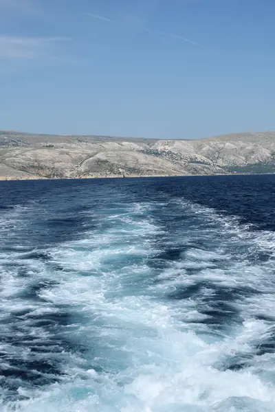 Köpüklü tekne derin mavi okyanus sularında iz bırakıyor, uzaktaki dağlık bir adaya doğru gidiyor..