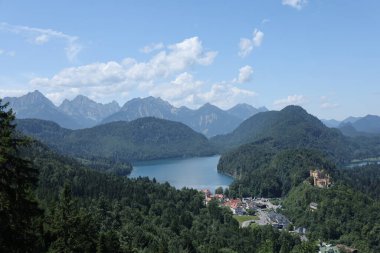 Sakin bir dağ gölü, yamaçta bir kale ve kabarık bulutların altında ormanla kaplı tepeler barındıran çarpıcı bir manzara..