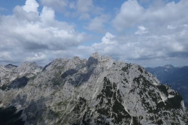 Ufuk boyunca uzanan bir dağ silsilesinin sarp ve dramatik zirveleri bulutlarla dolu dinamik bir gökyüzü altında.