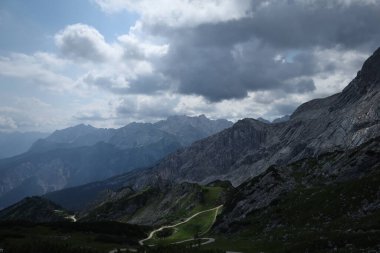Dolambaçlı bir toprak yol yeşil bir dağ vadisine çıkar. Etrafı katmanlı bulutlarla dolu bir gökyüzünün altındaki yüksek uçurumlarla çevrilidir. Vahşi doğa ve yolun güzel bir karışımı..