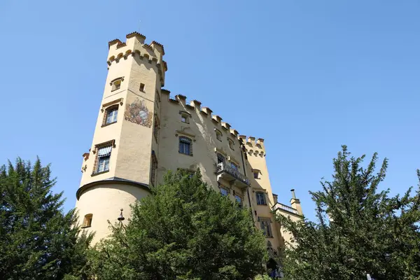 Açık mavi gökyüzünün altında dekoratif kuleleri ve ağaçları olan ortaçağdan kalma sarı bir kalenin kıvrımlı bir köşesi..