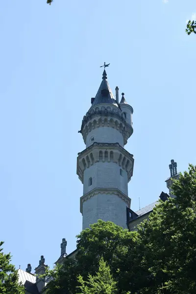 Yeşil ağaçlar ve mavi gökyüzüyle çevrili masal gibi bir kaleden yuvarlak bir taş kulenin dikey görüntüsü.