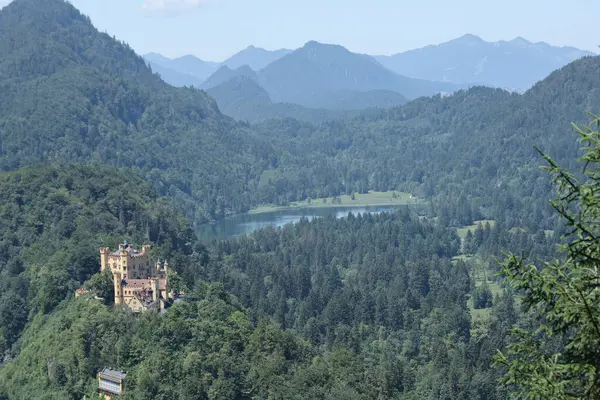 Bavyera dağlarıyla çevrili bir orman tepesine tünemiş görkemli sarı bir kale ve yaz gökyüzünün altında sakin bir göl..