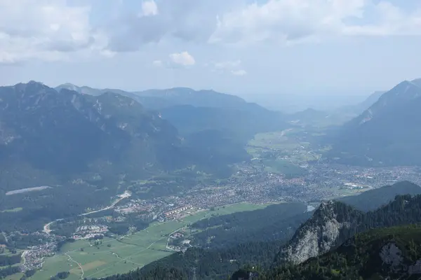 Parçalı bulutlu bir gökyüzünün altında ormanlık dağların arasına yerleşmiş bir kasaba olan bir Alp Vadisi 'nin geniş açılı görüntüsü. Sahne, doğal ve insan manzaraları arasındaki uyumu gösteriyor..