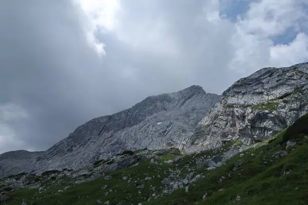 Yoğun fırtınalı bulutların arasından süzülen dar bir güneş ışığıyla aydınlatılan engebeli bir dağ manzarası..