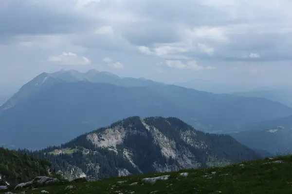 Uzak dağ sırtı katmanları sisli ufukta kaybolur, ön planda yeşil orman yamaçları vardır..