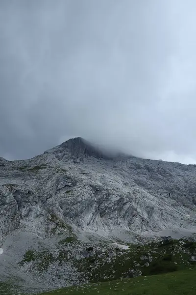 Yükselen bir dağın dikey fotoğrafı, zirvesi kalın bir sis ile gizlenmiş, engebeli uçurum dokularını ve aşağıdaki yeşilliği aydınlatan..