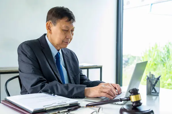 Attorney or lawyer in formal attire is sitting at desk with paper files and important documents and is working by using laptop to viewimportant information of cases to be brought to the courtroom. Law, justice concept.