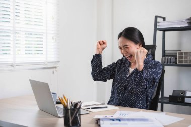 Genç iş kadını veya şirket çalışanı masasında oturuyor ve çalışmalarının tamamlanması, satış büyümesi veya başarılı projesinde başarısını kutluyor. Ellerini kaldırdı ve heyecanla gülümsedi..