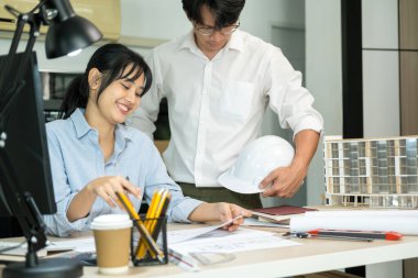 İki kadın ve erkek mühendis veya mimar çalışma masasında inşaat modeli ve ofis işyerindeki diğer ekipmanlarla birlikte çalışıyor. Erkek bir iş arkadaşı konuşurken inşaat projesinin planlarını, planlarını ve planlarını ele alıyor ve tartışıyorlar.