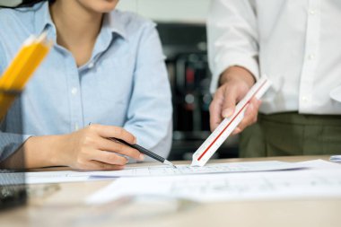 İki erkek ve kadın mühendis veya mimar çalışma masasında kağıt işleri ve ofis işyerindeki diğer ekipmanlarla birlikte çalışarak bir cetveli işaret ediyor, inşaat projesinin planları hakkında plan yapıyor ve tartışıyorlar..